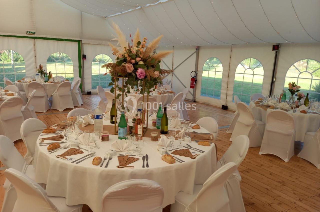 Salle de réception sous chapiteau avec tables rondes dressées, nappes blanches et décoration florale centrale.