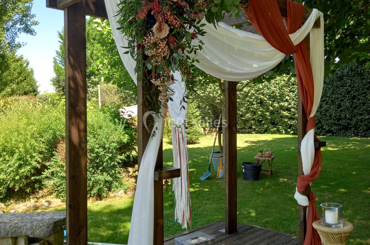 Arche en bois décorée de fleurs, tissus blancs et rouges, installée dans un jardin verdoyant.