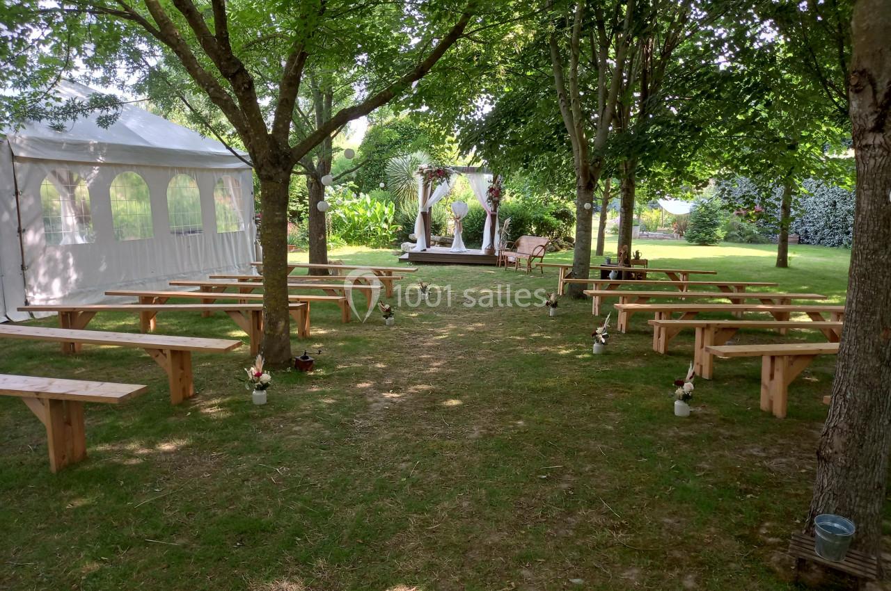 Espace extérieur aménagé pour une cérémonie, avec bancs en bois, tonnelle décorée et tente blanche sous des arbres.