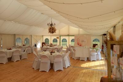 Deux balais déguisés en mariés avec robe et costume, posés devant une tente blanche et un cadre décoratif.