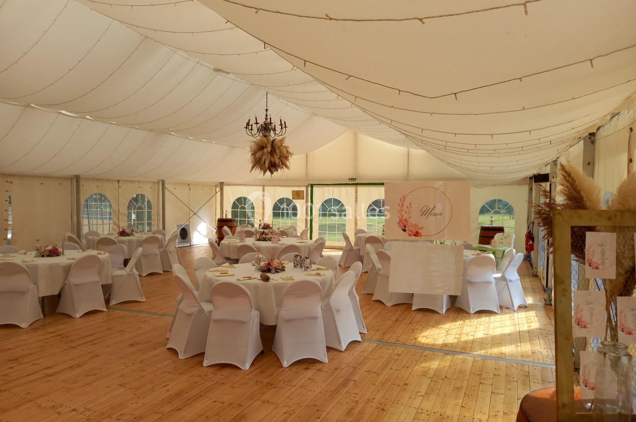 Salle de réception sous une tente, décorée avec des tables rondes, nappes blanches et guirlandes lumineuses suspendues.