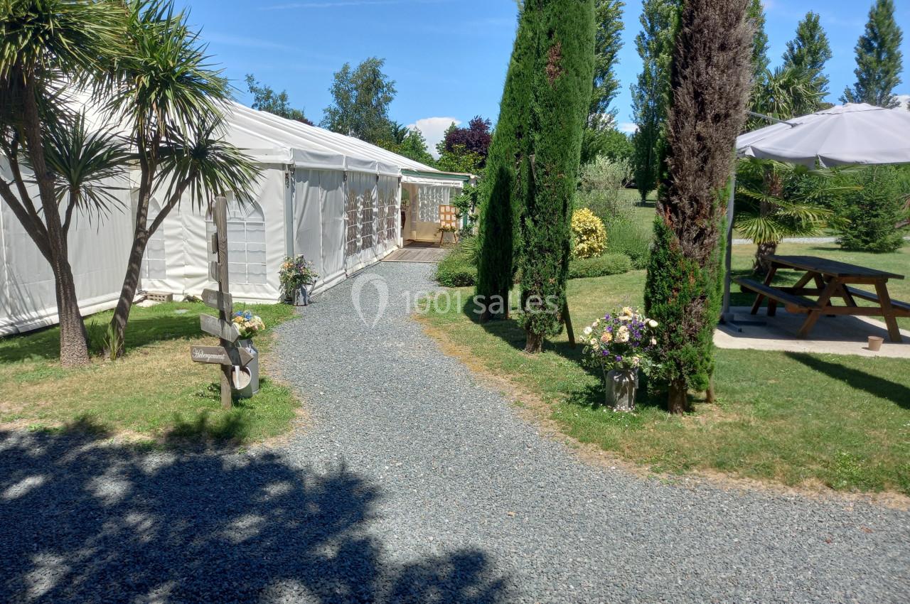 Allée gravillonnée bordée de verdure menant à une grande tente blanche, avec des tables de pique-nique à proximité.