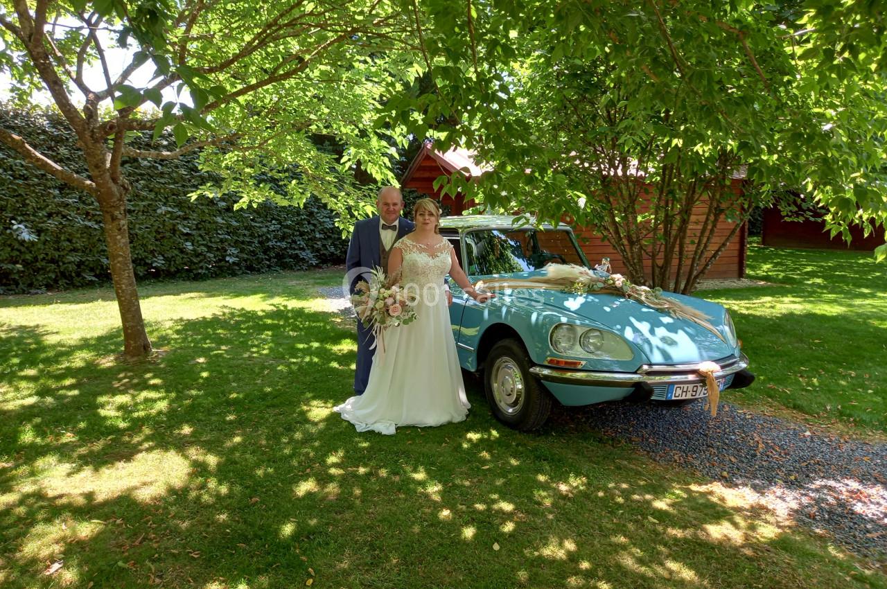 Un couple en tenue de mariage pose devant une voiture ancienne bleu clair dans un jardin ombragé.