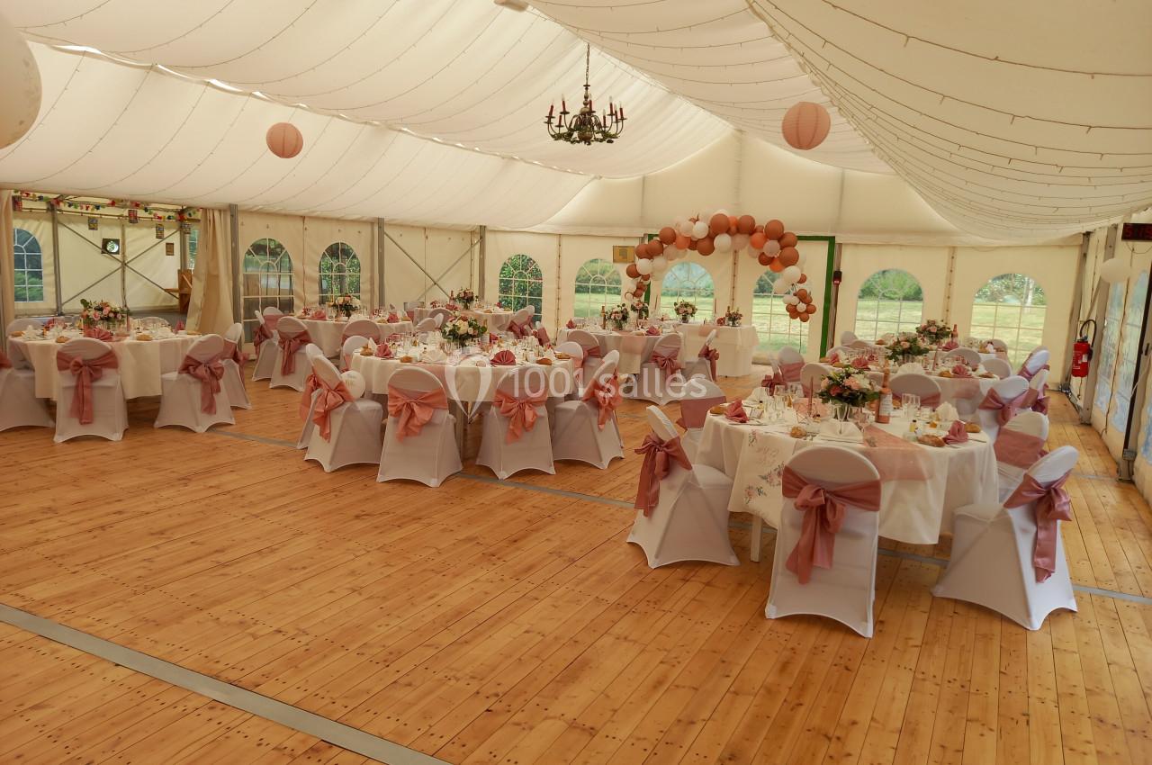 Salle de réception décorée pour un mariage, avec tables dressées, chaises ornées de nœuds roses et guirlandes lumineuses.