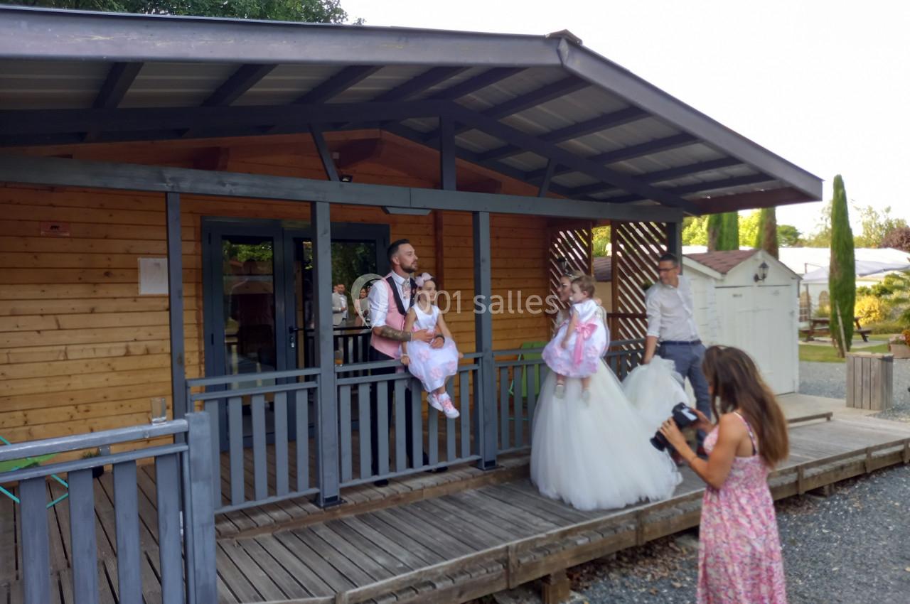 Un homme pose avec deux enfants en tenue de cérémonie sur la terrasse d'un chalet en bois, une photographe les prend en…
