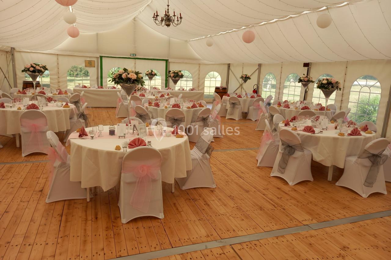 Salle de réception sous chapiteau avec tables rondes décorées de nappes blanches et nœuds roses sur les chaises.