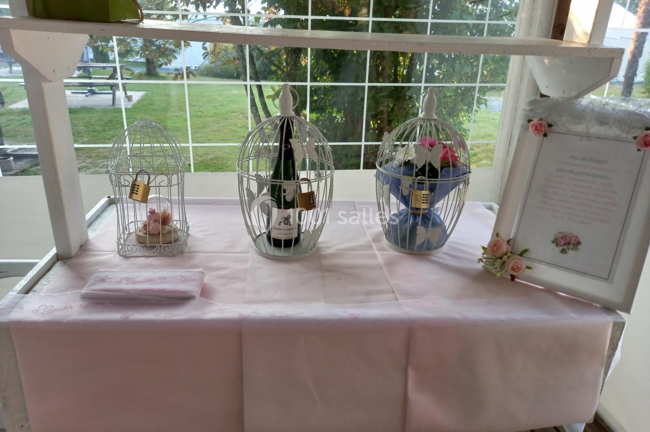 Trois cages décoratives sur une table rose, contenant une bouteille, des bougies et des fleurs, avec un cadre à droite.