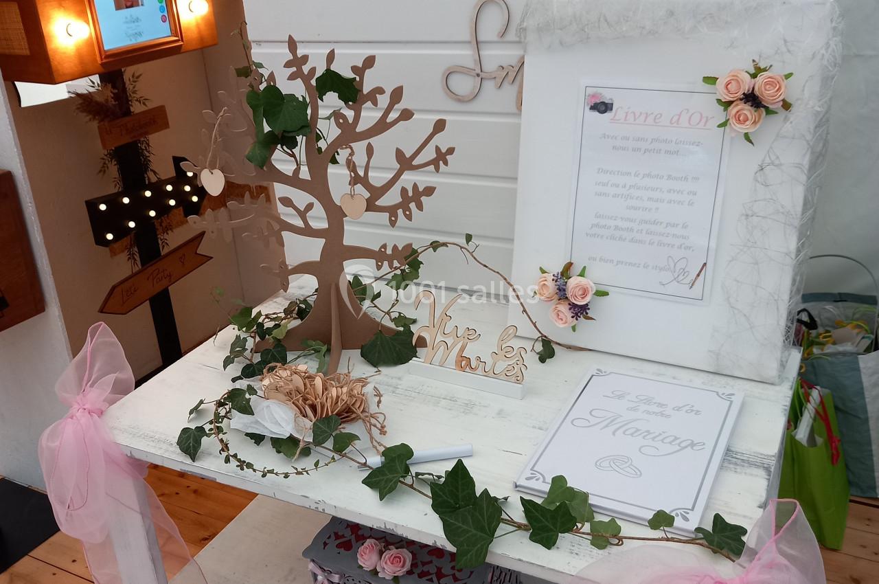 Décoration de mariage avec un arbre à vœux, des roses, des cadres et une boîte sur une table blanche ornée de rubans roses.