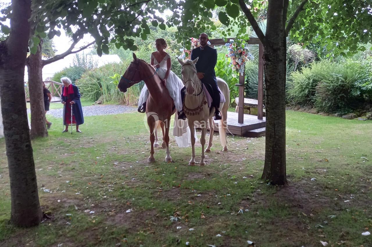 Un couple en tenue de mariage est assis sur des chevaux dans un jardin, entouré d'arbres et de verdure.