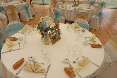 Deux balais déguisés en mariés avec robe et costume, posés devant une tente blanche et un cadre décoratif.