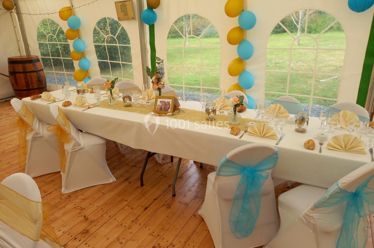 Table décorée pour un événement dans une tente, avec nappes blanches, chaises ornées de rubans et ballons colorés.