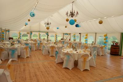 Deux balais déguisés en mariés avec robe et costume, posés devant une tente blanche et un cadre décoratif.