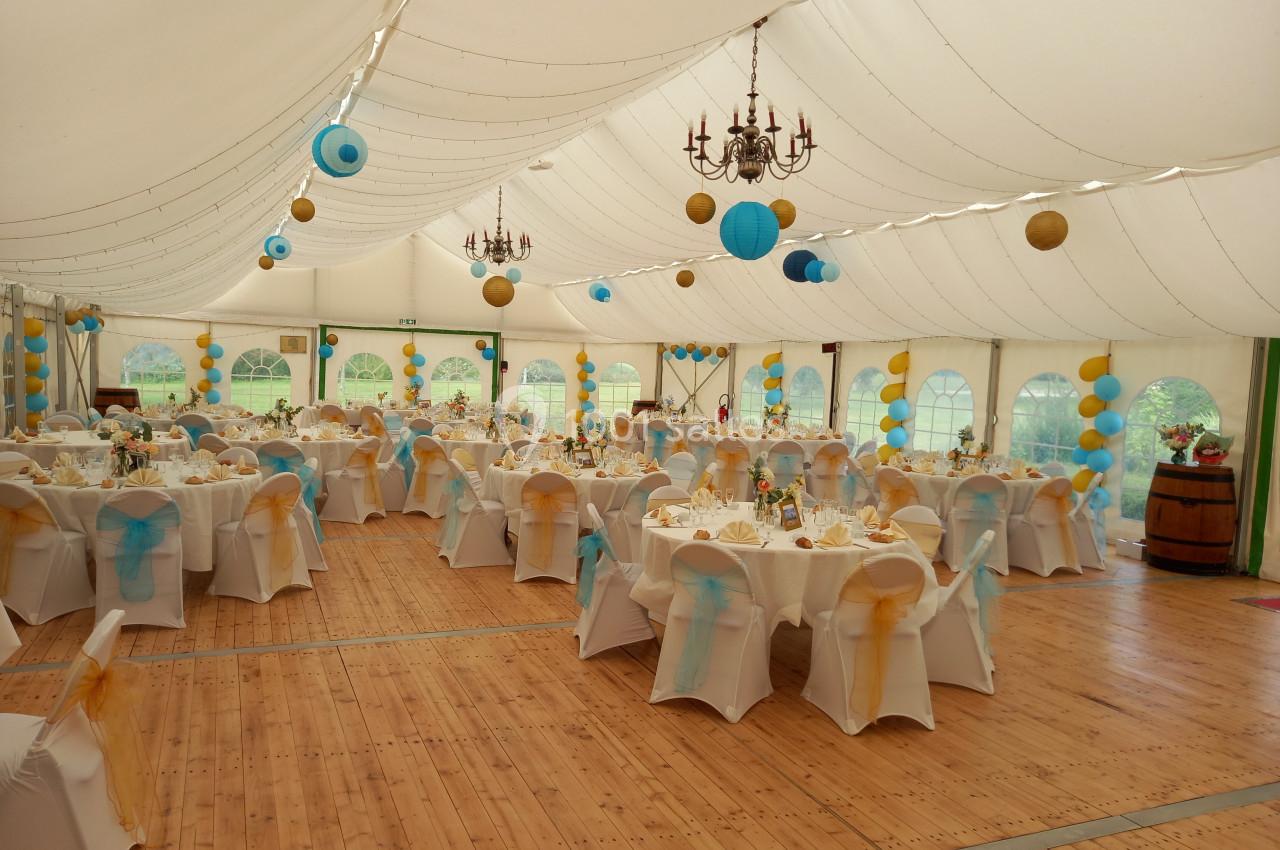 Salle de réception sous chapiteau avec tables décorées en blanc, bleu et jaune, éclairée par des lustres et des guirlandes…