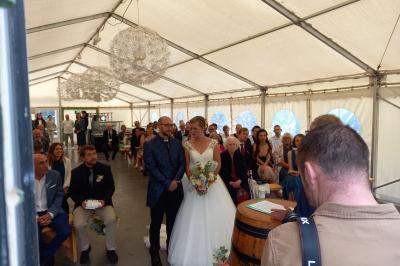 Deux balais déguisés en mariés avec robe et costume, posés devant une tente blanche et un cadre décoratif.