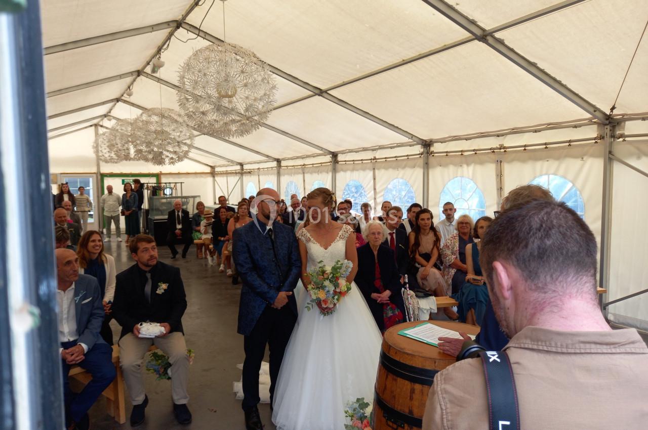 Un couple de mariés debout devant un officiant dans une tente, entourés d'invités assis.