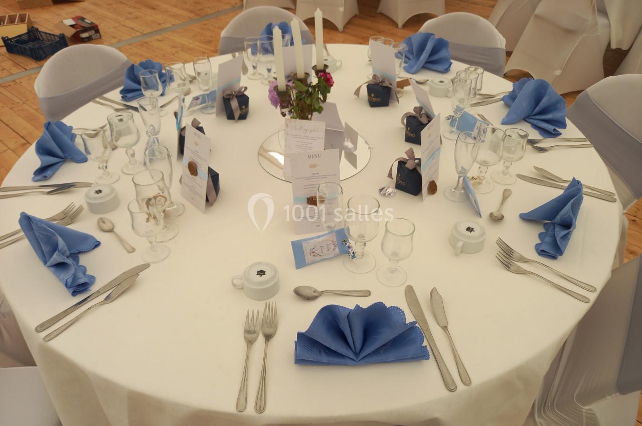 Table ronde dressée avec nappes blanches, serviettes bleues pliées, couverts, verres, menus et centre de table floral.