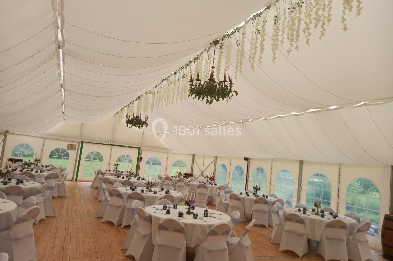 Intérieur d'une tente de réception avec tables rondes dressées, chaises blanches et décorations suspendues.