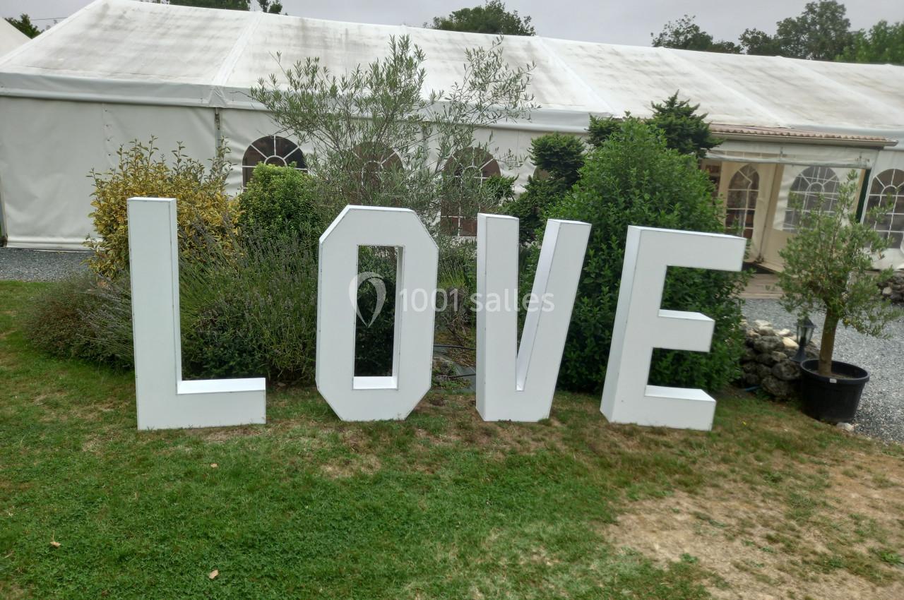 Lettres blanches formant le mot ’LOVE’ disposées sur une pelouse devant une grande tente blanche.
