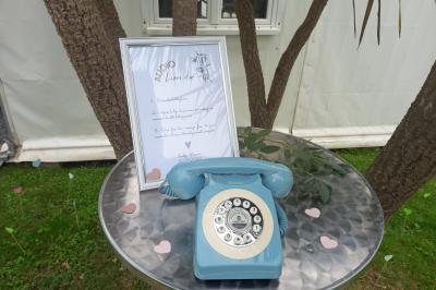 Deux balais déguisés en mariés avec robe et costume, posés devant une tente blanche et un cadre décoratif.