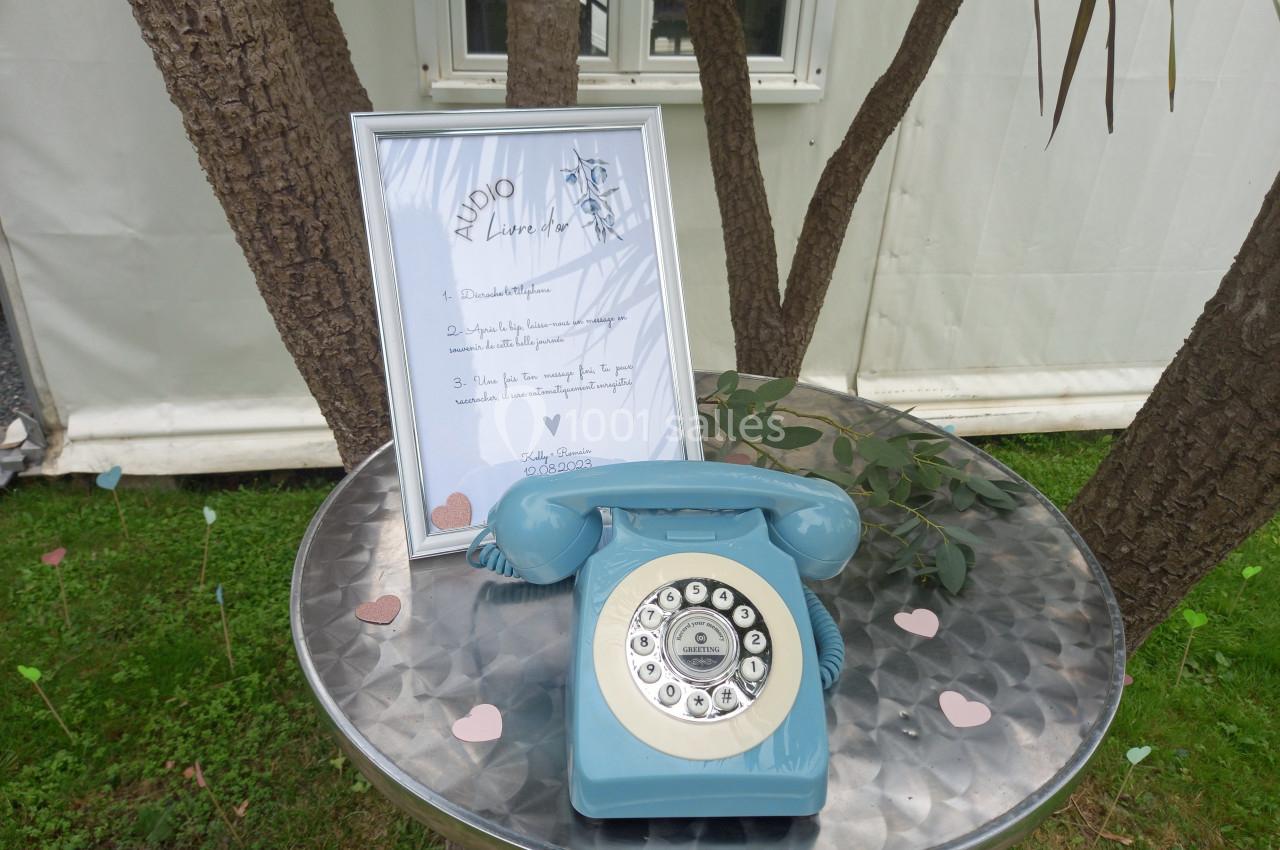 Téléphone rétro bleu posé sur une table en métal avec un cadre contenant un texte, devant un arbre et une tente blanche.