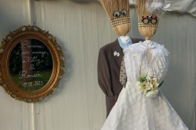 Deux balais déguisés en mariés avec robe et costume, posés devant une tente blanche et un cadre décoratif.