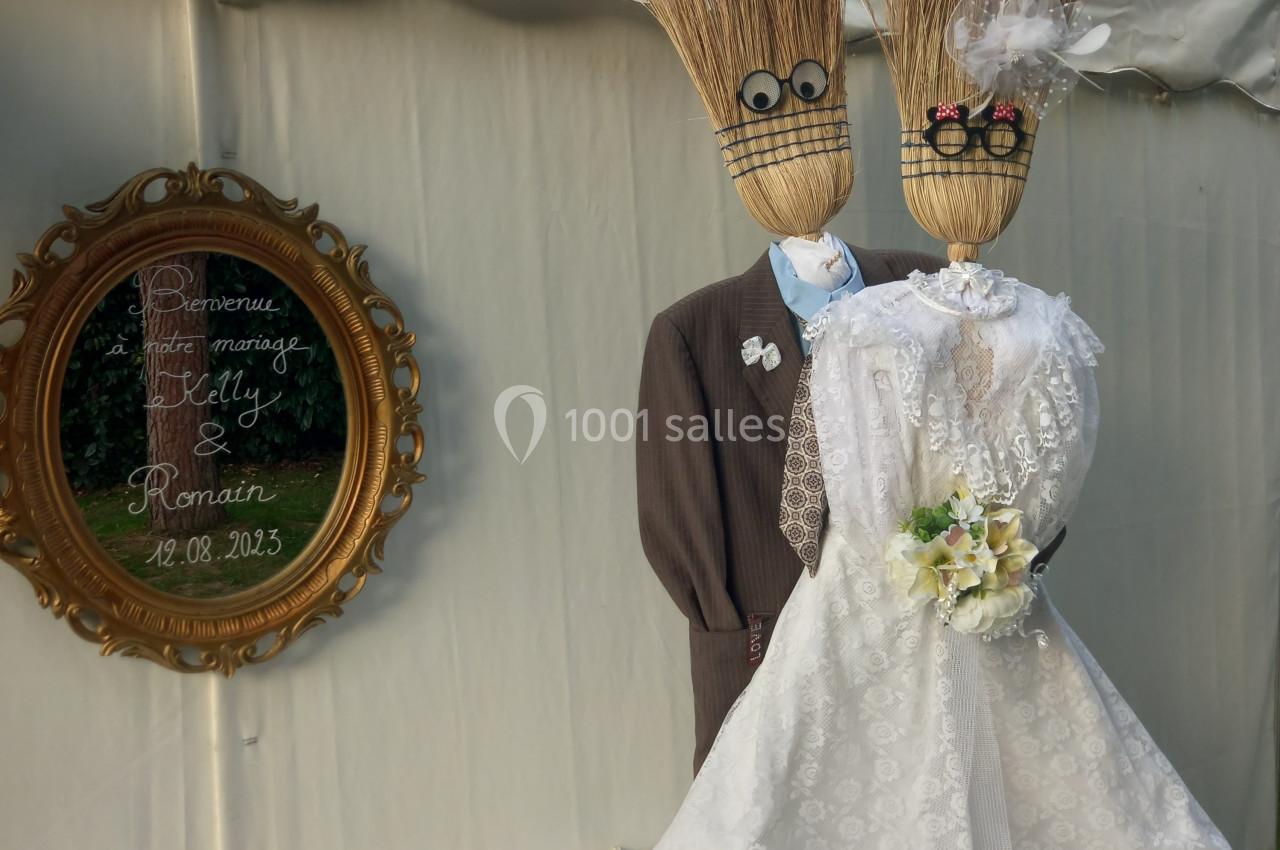 Deux balais déguisés en mariés avec robe et costume, posés devant une tente blanche et un cadre décoratif.
