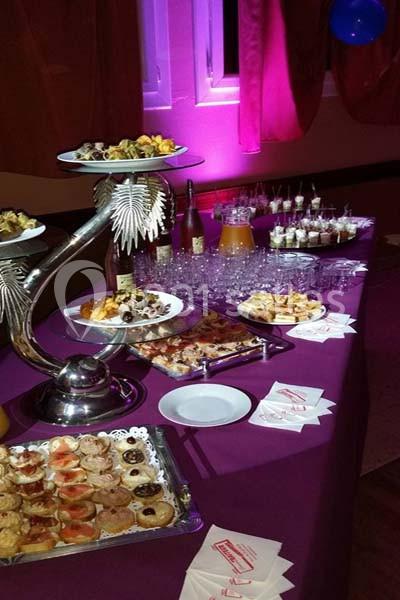 Buffet avec divers plats, verrines et boissons disposés sur une table violette dans une salle éclairée en rose.