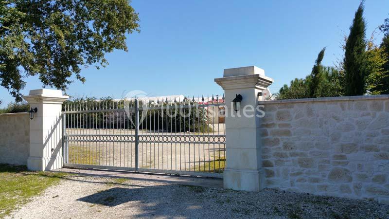 Portail métallique gris ouvert, encadré par des piliers en pierre, donnant sur une cour gravillonnée avec végétation en…