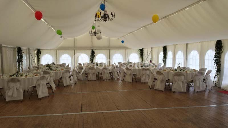 Salle de réception sous une tente blanche, décorée de ballons colorés et de tables dressées avec nappes blanches.