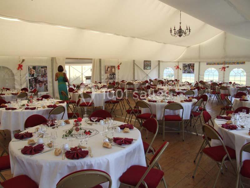 Salle de réception sous chapiteau, tables rondes dressées avec nappes blanches, serviettes rouges et décorations.