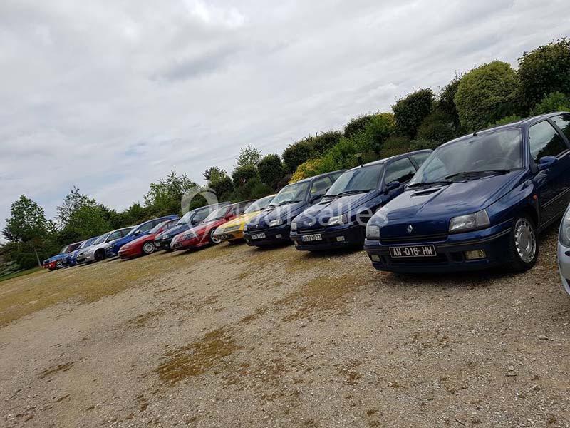 Rangée de voitures anciennes et modernes stationnées sur un parking en gravier, entouré de verdure.