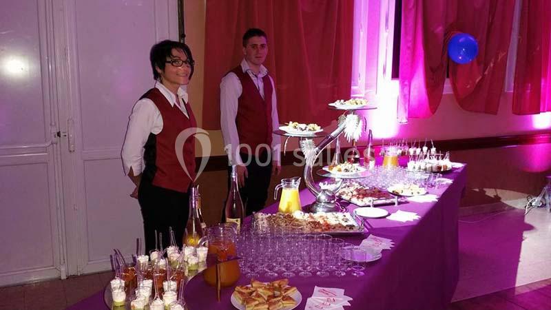 Deux serveurs en tenue de service devant une table garnie de boissons et d'amuse-bouches dans une salle éclairée.