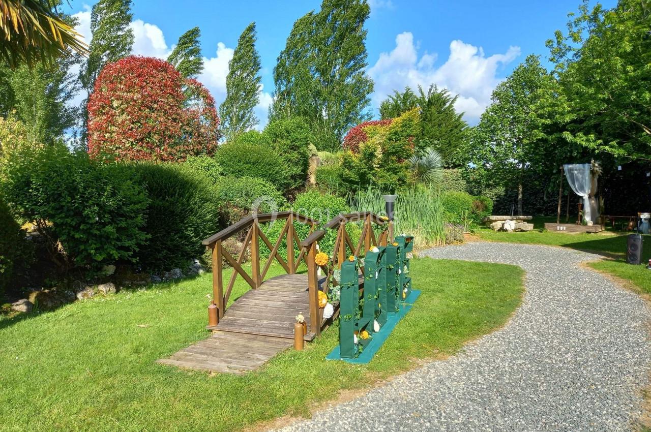 Petit pont en bois traversant une pelouse verdoyante, entouré d'arbustes et d'arbres sous un ciel bleu.