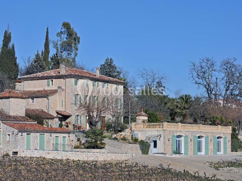 Bâtisse en pierre avec volets verts entourée de végétation, située sur un terrain ensoleillé avec vignes au premier plan.