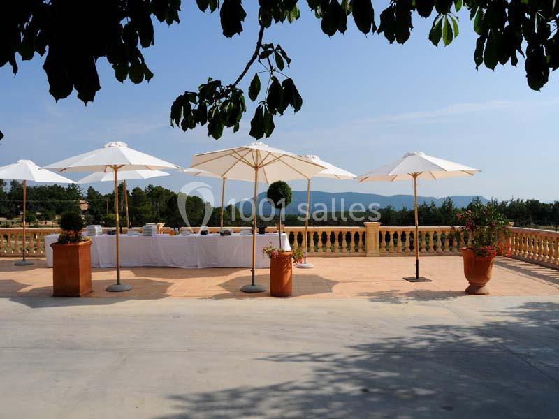 Terrasse ensoleillée avec parasols blancs, table dressée et vue sur un paysage vallonné sous un ciel dégagé.