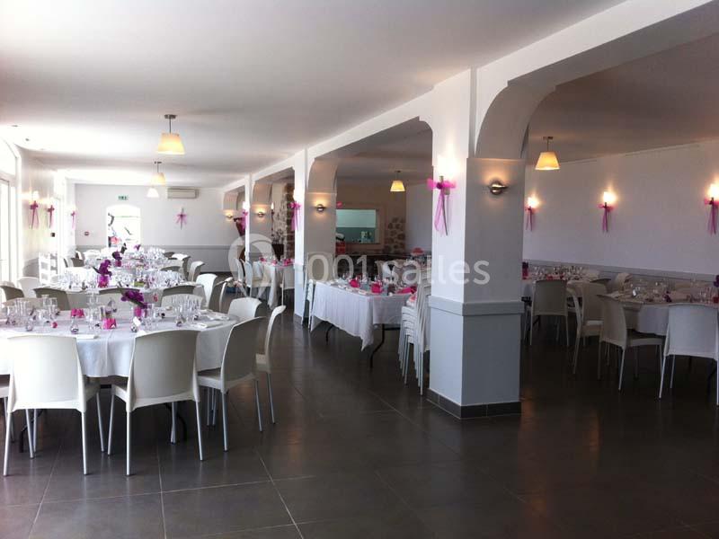 Salle de réception lumineuse avec des tables dressées, décorations roses et chaises blanches.