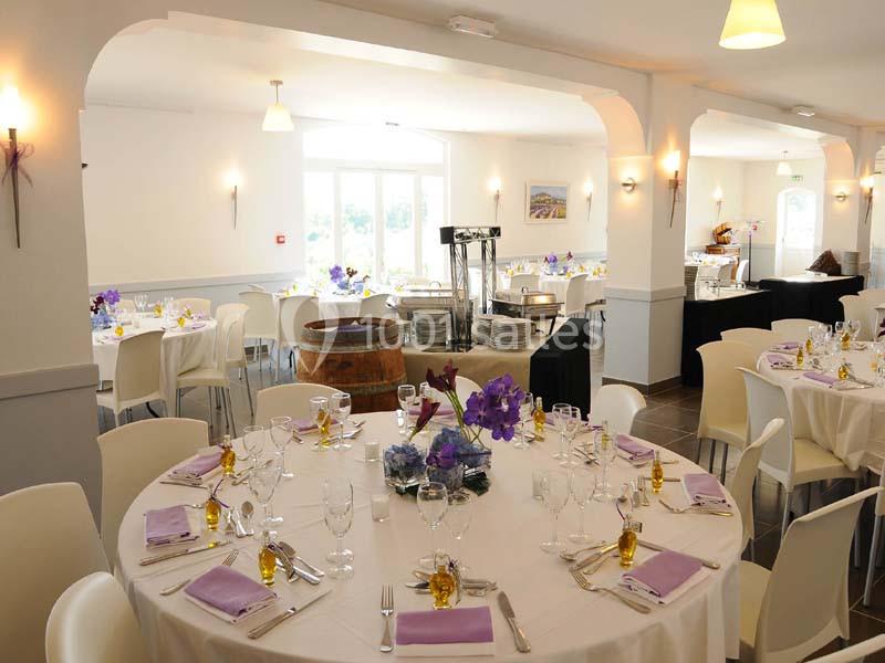 Salle de réception lumineuse avec tables rondes dressées, décorées de fleurs violettes et serviettes assorties.