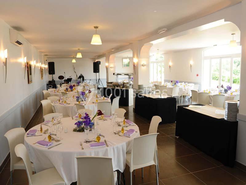Salle de réception lumineuse avec tables rondes dressées, décorées de fleurs violettes, et buffet en arrière-plan.