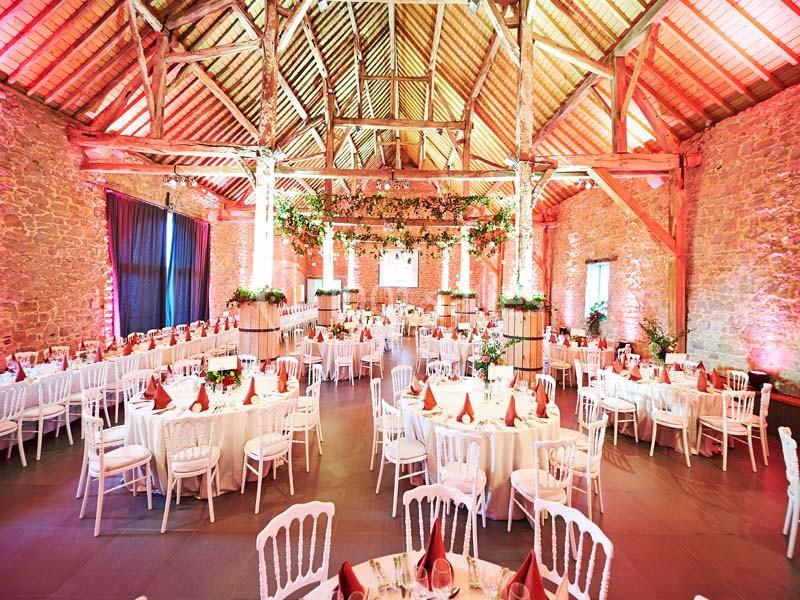 Salle de réception en pierre avec poutres apparentes, tables rondes dressées et décorations florales suspendues.