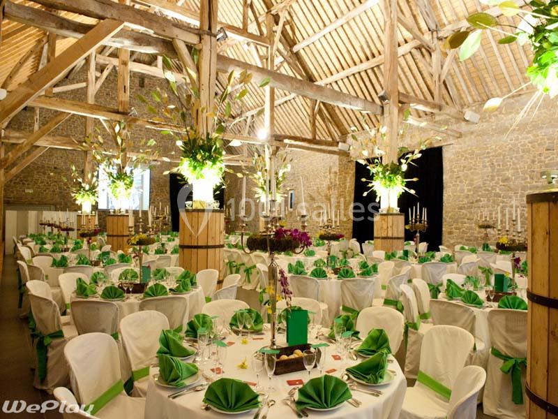 Salle de réception décorée avec des tables rondes, nappes blanches, serviettes vertes et arrangements floraux suspendus.