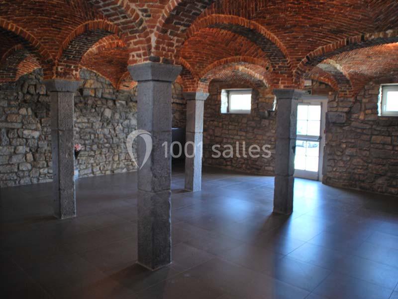Salle voûtée en briques avec piliers en pierre, sol en carrelage sombre et lumière naturelle entrant par des fenêtres.
