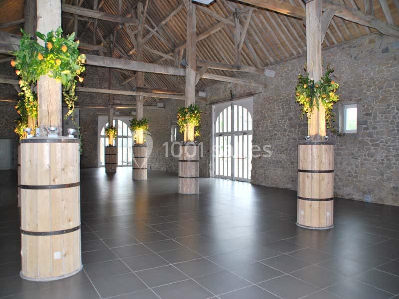 Salle spacieuse avec poutres en bois, piliers décorés de plantes et grandes fenêtres laissant entrer la lumière naturelle.