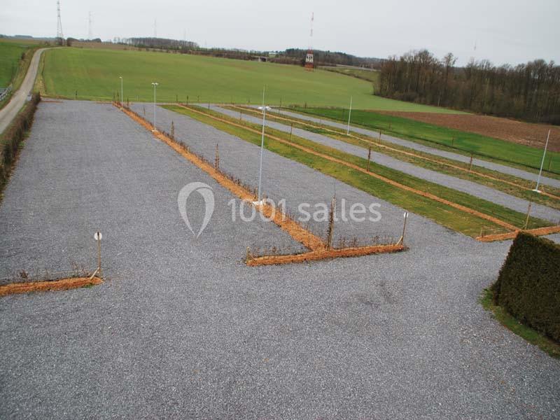 Parking en gravier vide, délimité par des haies et entouré de champs et d'arbres sous un ciel nuageux.