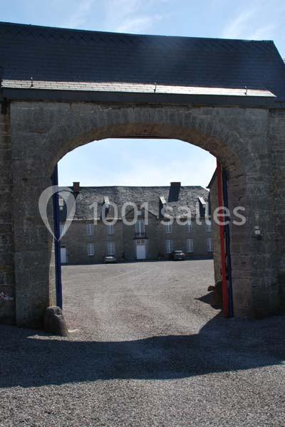 Vue d'une cour pavée à travers une arche en pierre, avec des bâtiments en arrière-plan sous un ciel dégagé.