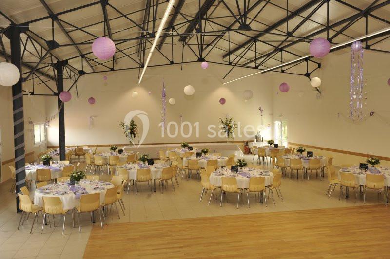 Salle de réception décorée avec des tables rondes, des chaises en bois clair et des suspensions violettes et blanches.