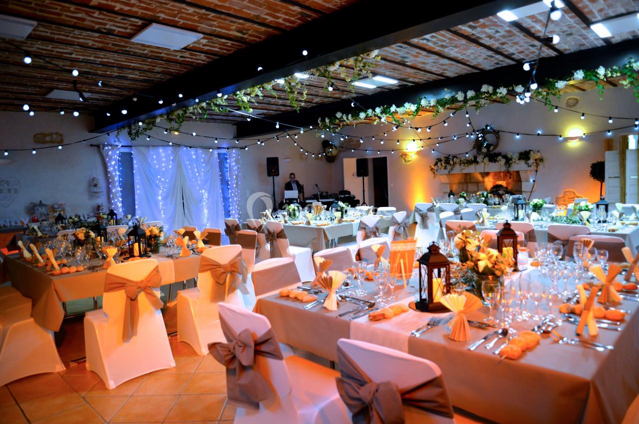 Salle de réception décorée pour un mariage avec tables dressées, chaises ornées de nœuds et guirlandes lumineuses suspendues.