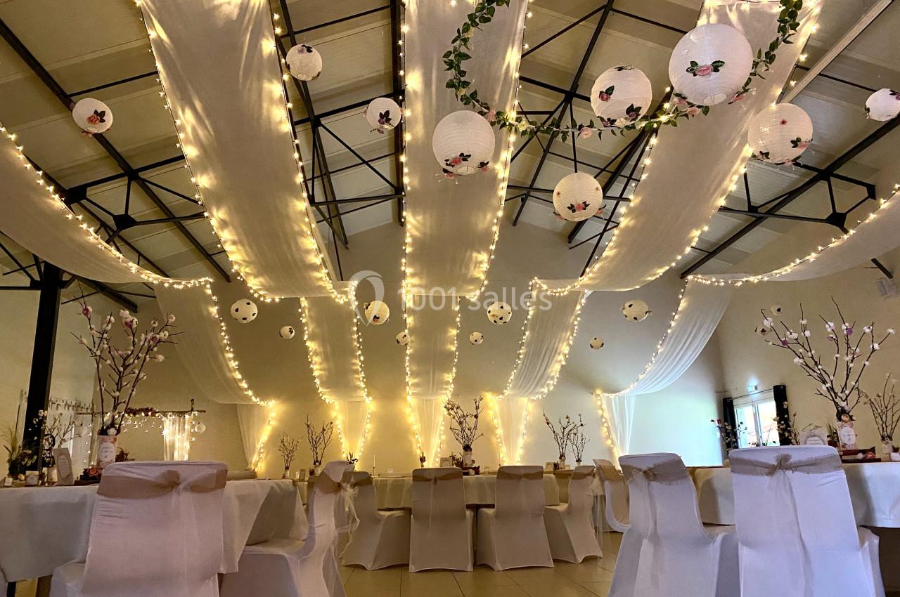 Salle décorée pour un événement avec guirlandes lumineuses, lanternes suspendues et chaises recouvertes de housses blanches.