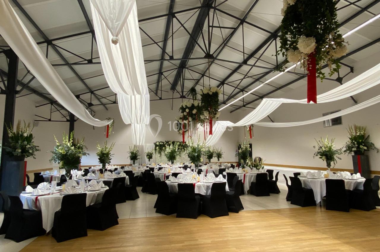 Salle de réception décorée avec des tables rondes dressées, nappes blanches, chaises noires et drapés suspendus.