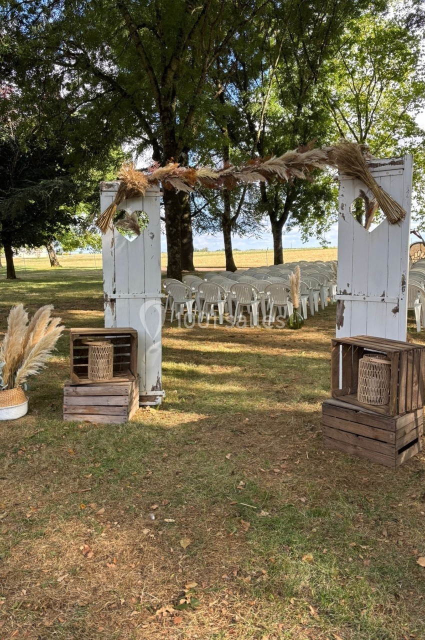 Arche en bois blanc décorée de fleurs séchées, entourée de chaises blanches disposées en extérieur sous des arbres.