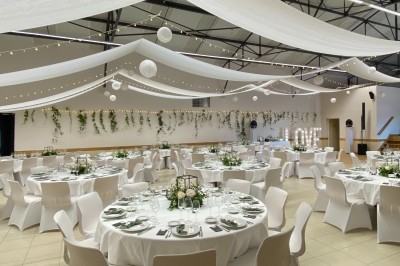 Salle avec des rangées de chaises en plastique beige face à une scène surélevée dans un espace lumineux.